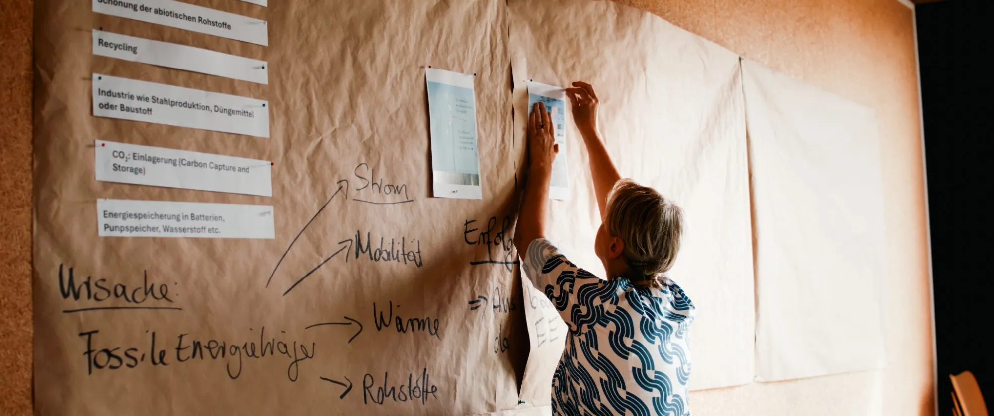 Eine Frau befestigt ein Schaubild zu erneuerbaren Energien an einer Papierwand, auf der „Ursache: Fossile Energieträger“ steht